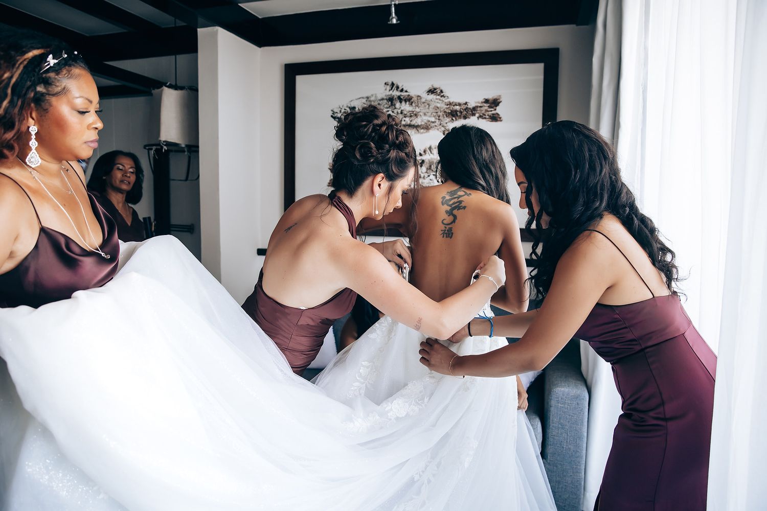 Wedding party members in burgundy dresses help adjust the bride's gown in a getting ready room.