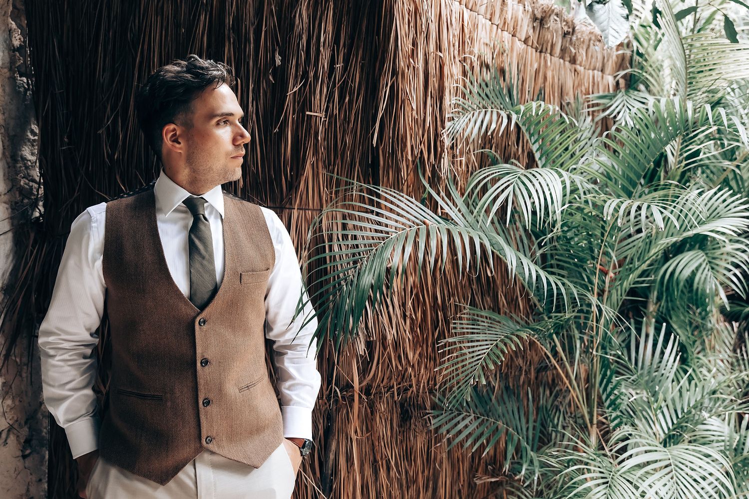 Wedding couple in elegant attire poses against a tropical backdrop with palm trees and thatched decor.