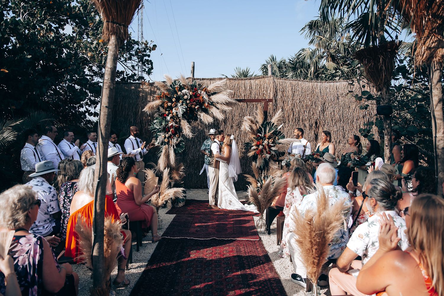 A group of people gathered outdoors at a social event with palm trees and decorative elements in the background.
