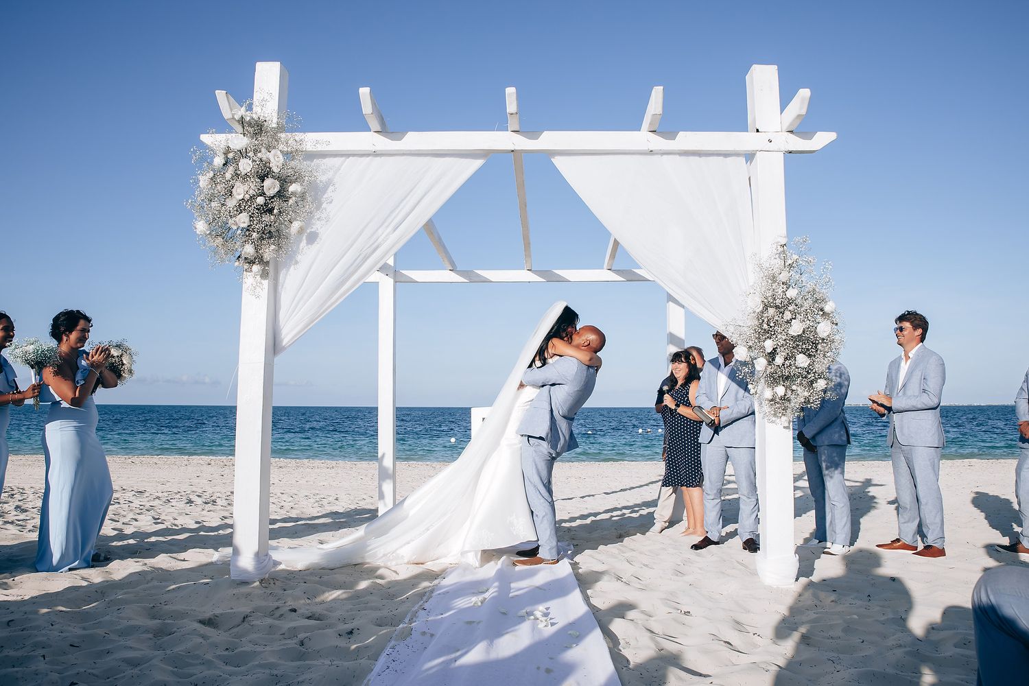 Beach wedding ceremony setup with white arch and chairs against blue ocean backdrop.