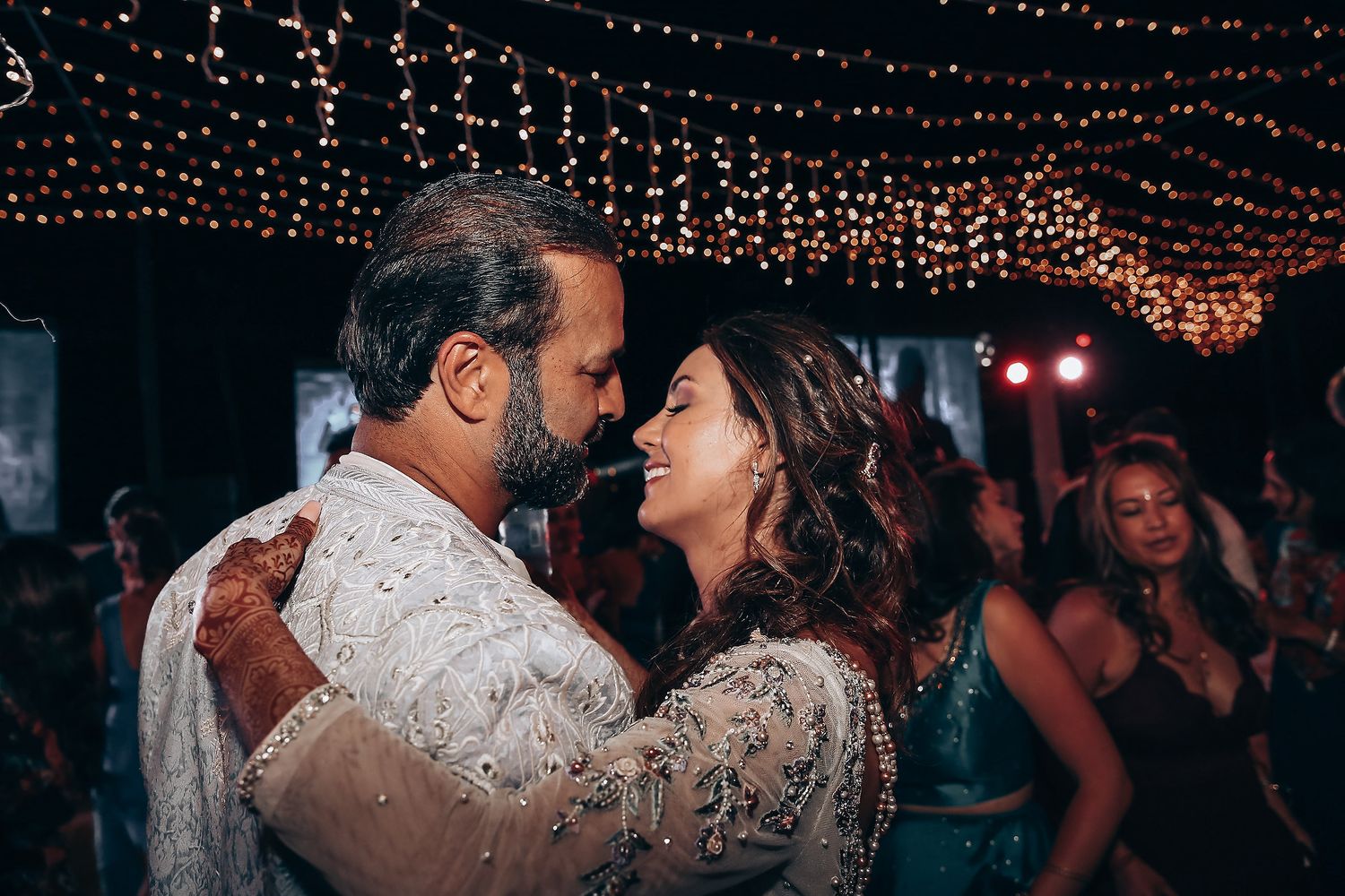Couple dancing together at an evening reception under twinkling string lights and chandeliers.