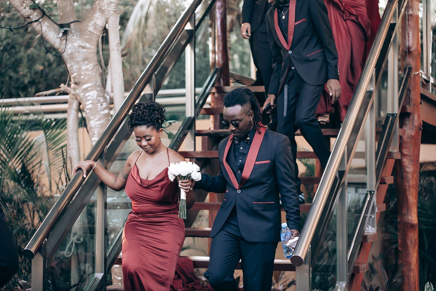 A couple in burgundy dress and navy suit share an intimate moment on outdoor stairs during a wedding photoshoot.