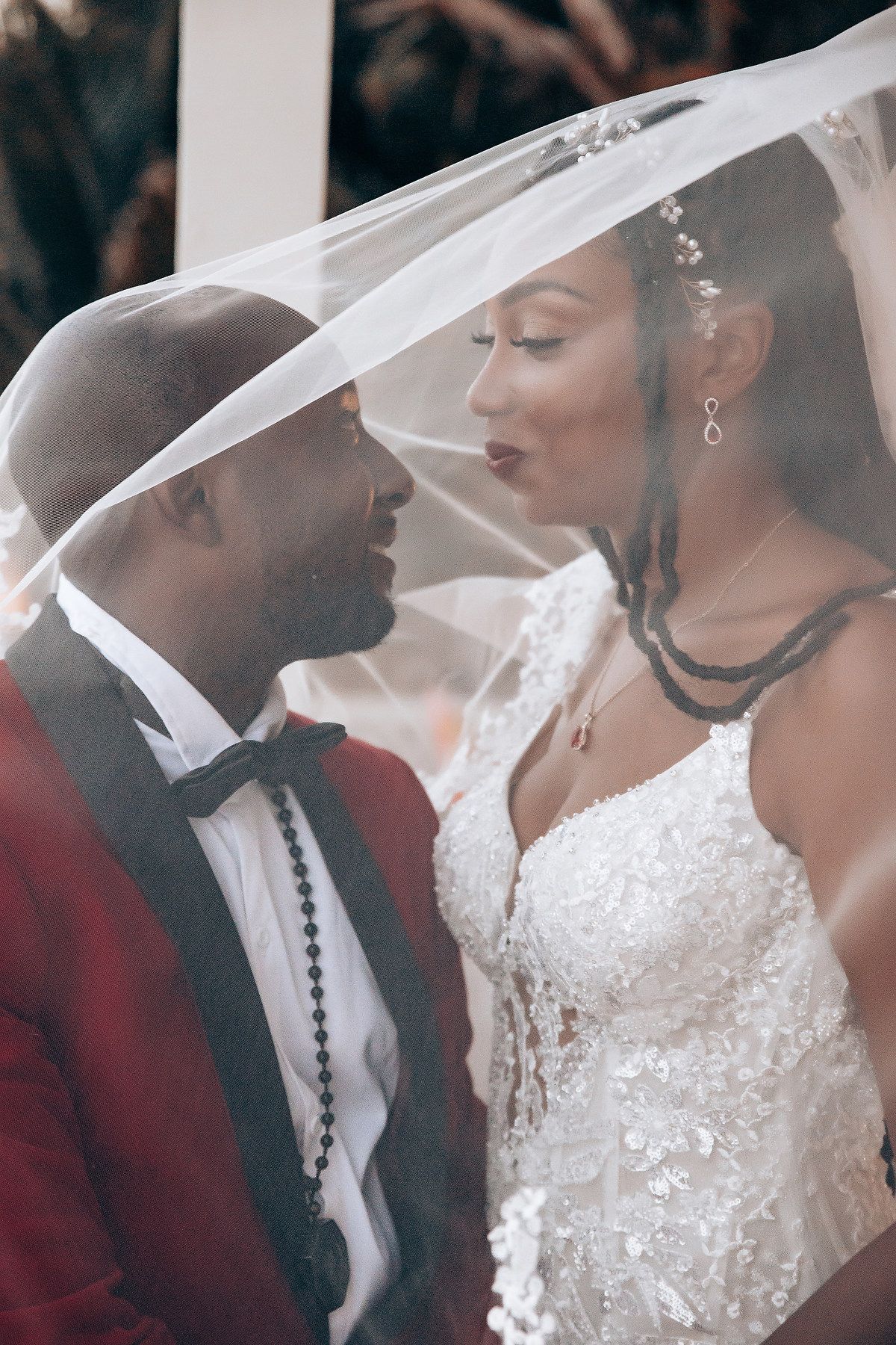 A romantic wedding moment captured through a delicate veil, with the groom in a burgundy tuxedo and bride in lace gown.