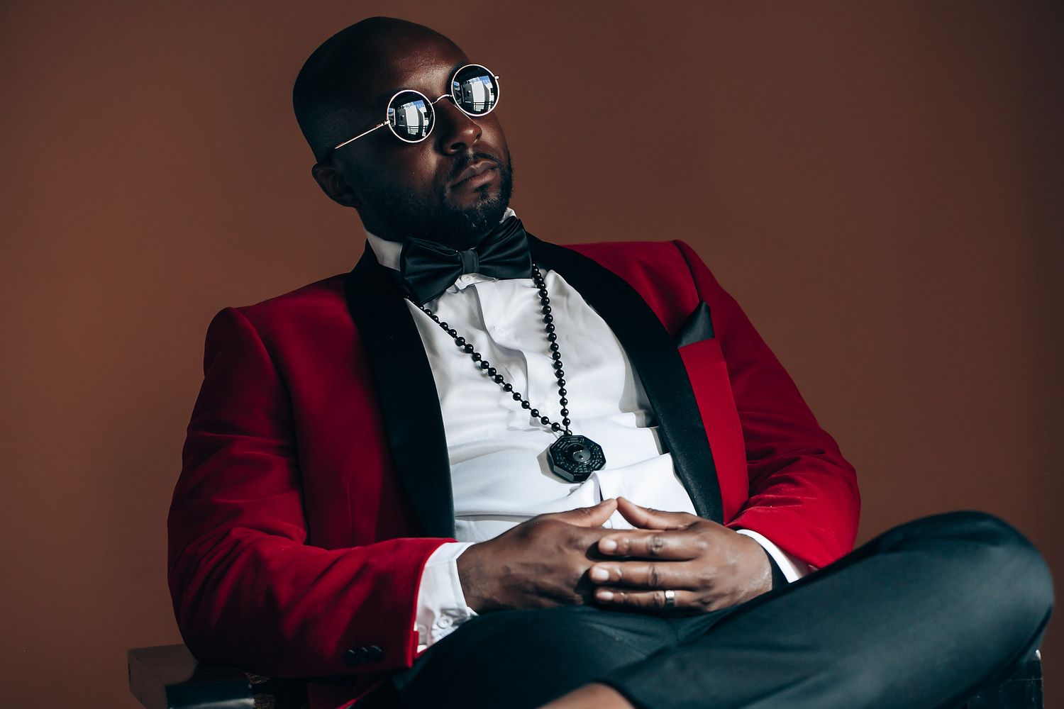 A person in a red velvet blazer and bowtie sits contemplatively wearing glasses and a necklace against a brown backdrop.