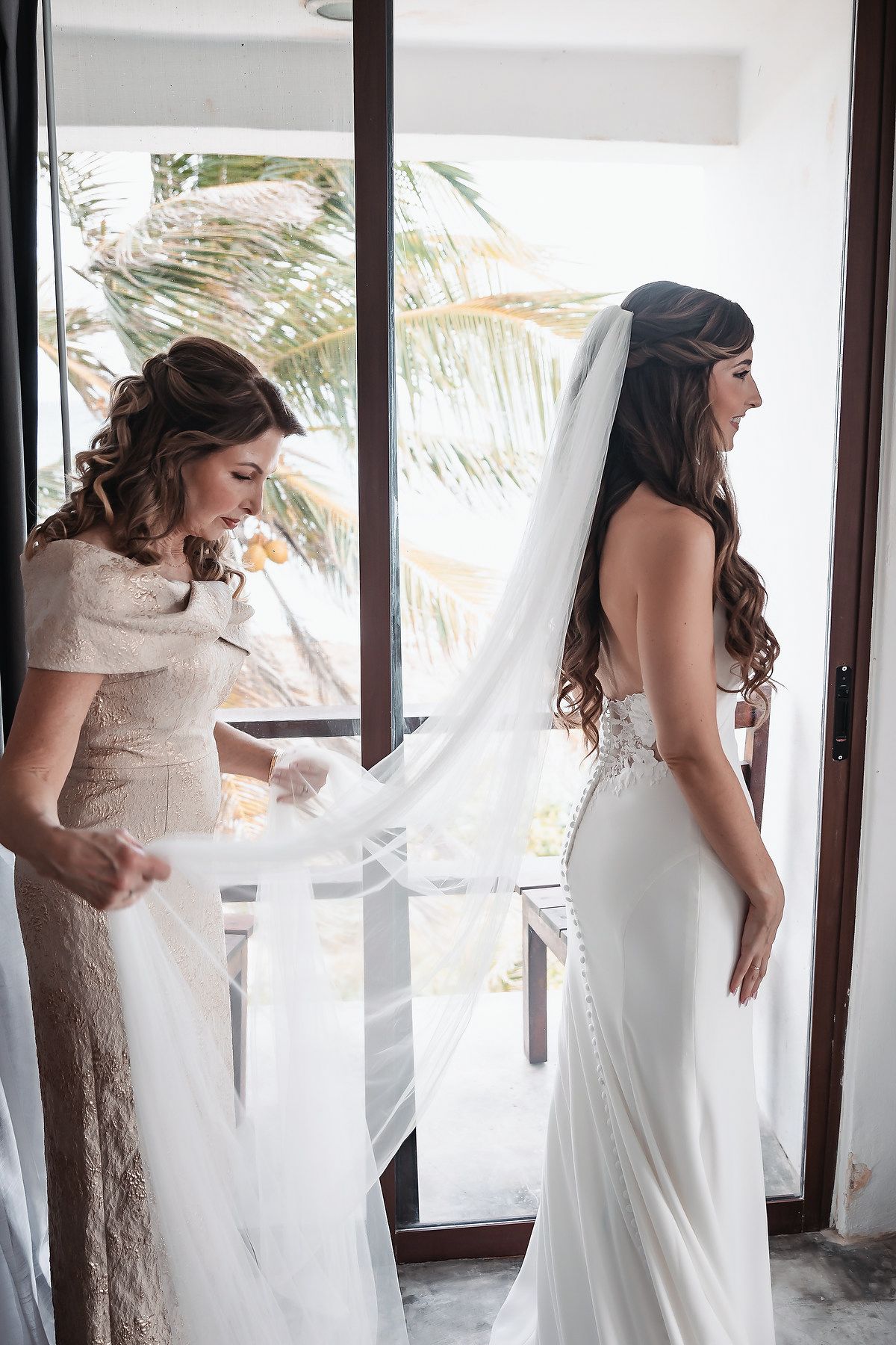 A bride in a white gown and veil stands in a doorway as a woman in a lace dress adjusts her veil.