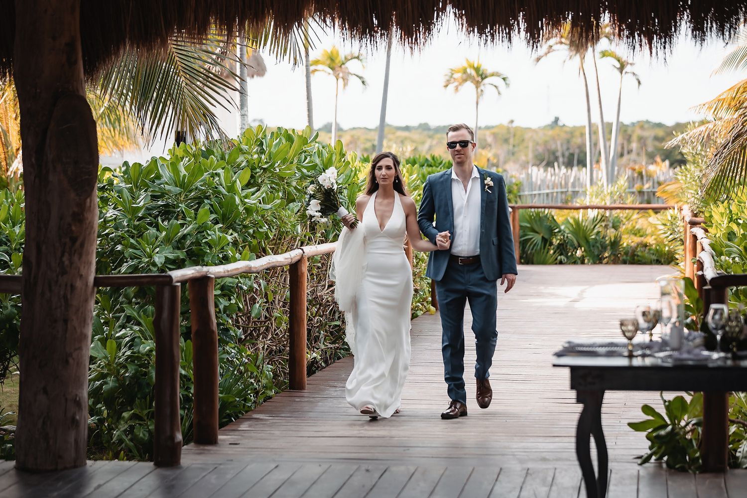 Bride and groom walking hand in hand on a tropical resort boardwalk surrounded by lush greenery and palm trees.