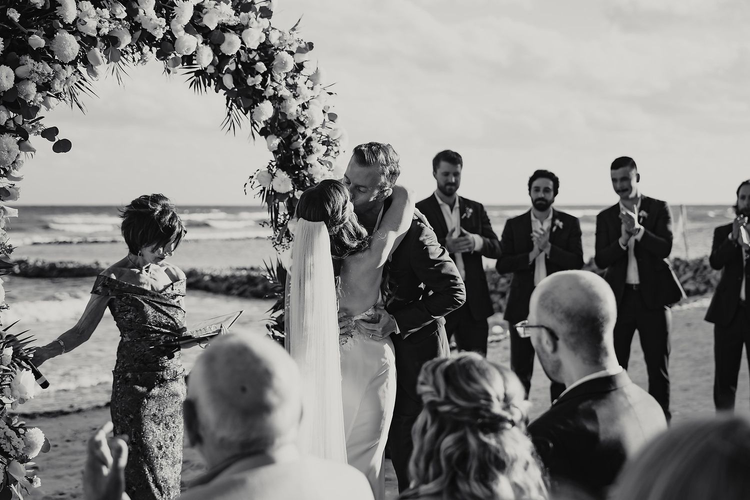 Bride and groom share first kiss under floral arch at beach wedding ceremony in black and white.