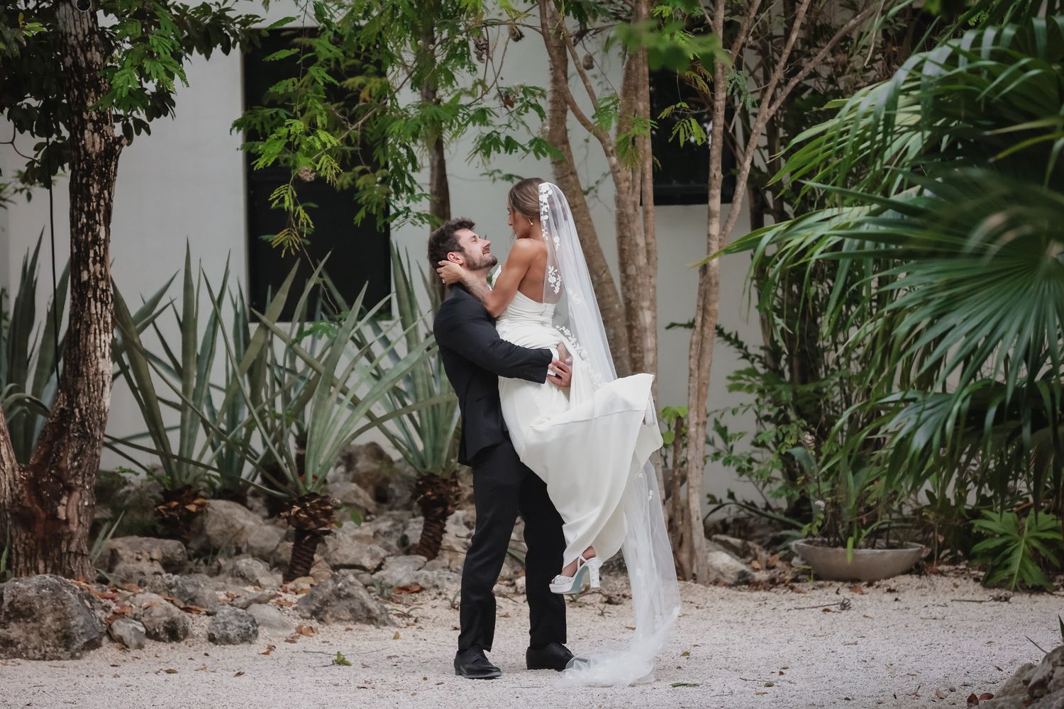 Groom lifts bride in white wedding gown and veil surrounded by tropical greenery outdoors.