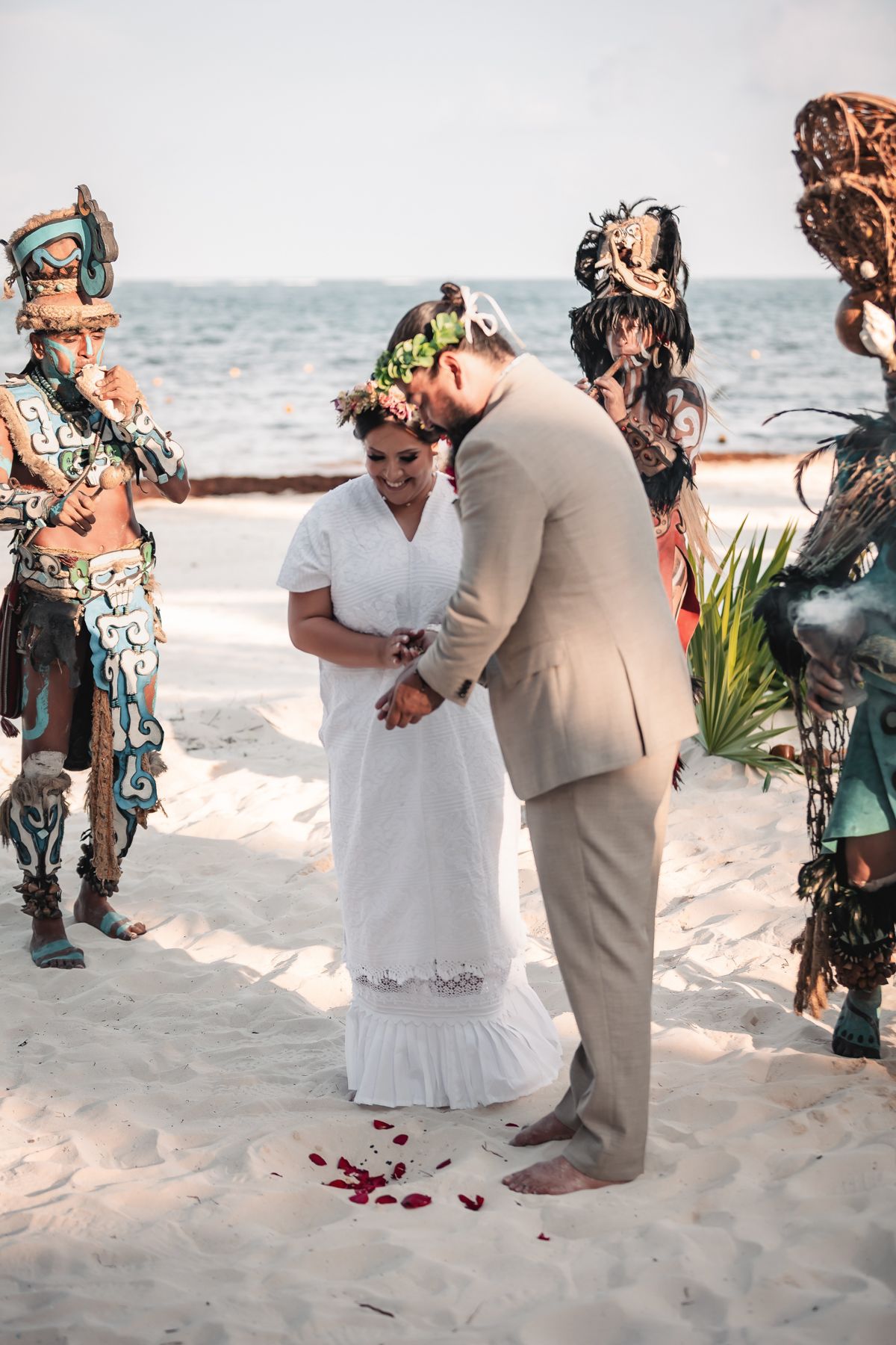 Couple exchanges vows on sandy beach surrounded by Mayan performers in traditional costumes during a destination wedding ceremony.