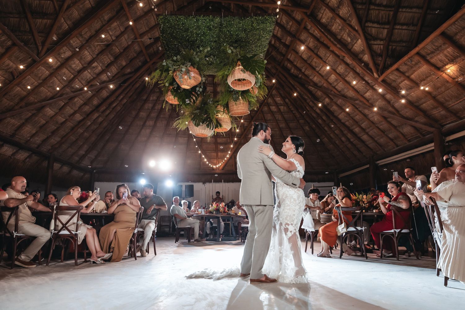 Bride and groom share first dance under floral chandelier in rustic barn wedding venue with string lights.