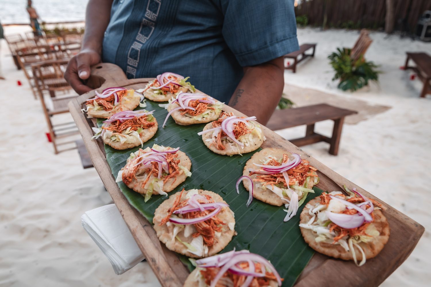Person holding a wooden board with mini flatbread appetizers topped with red onions and toppings on banana leaves at a beach.