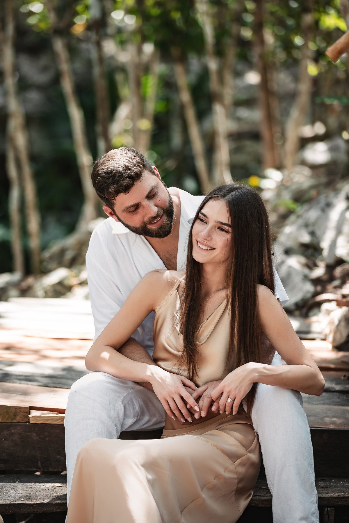 Happy couple sitting together outdoors, man embracing smiling woman with long hair in a sunlit forest setting.