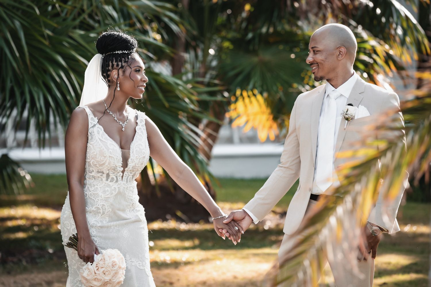 A Black bride and groom hold hands outdoors in wedding attire, surrounded by lush green palm trees.