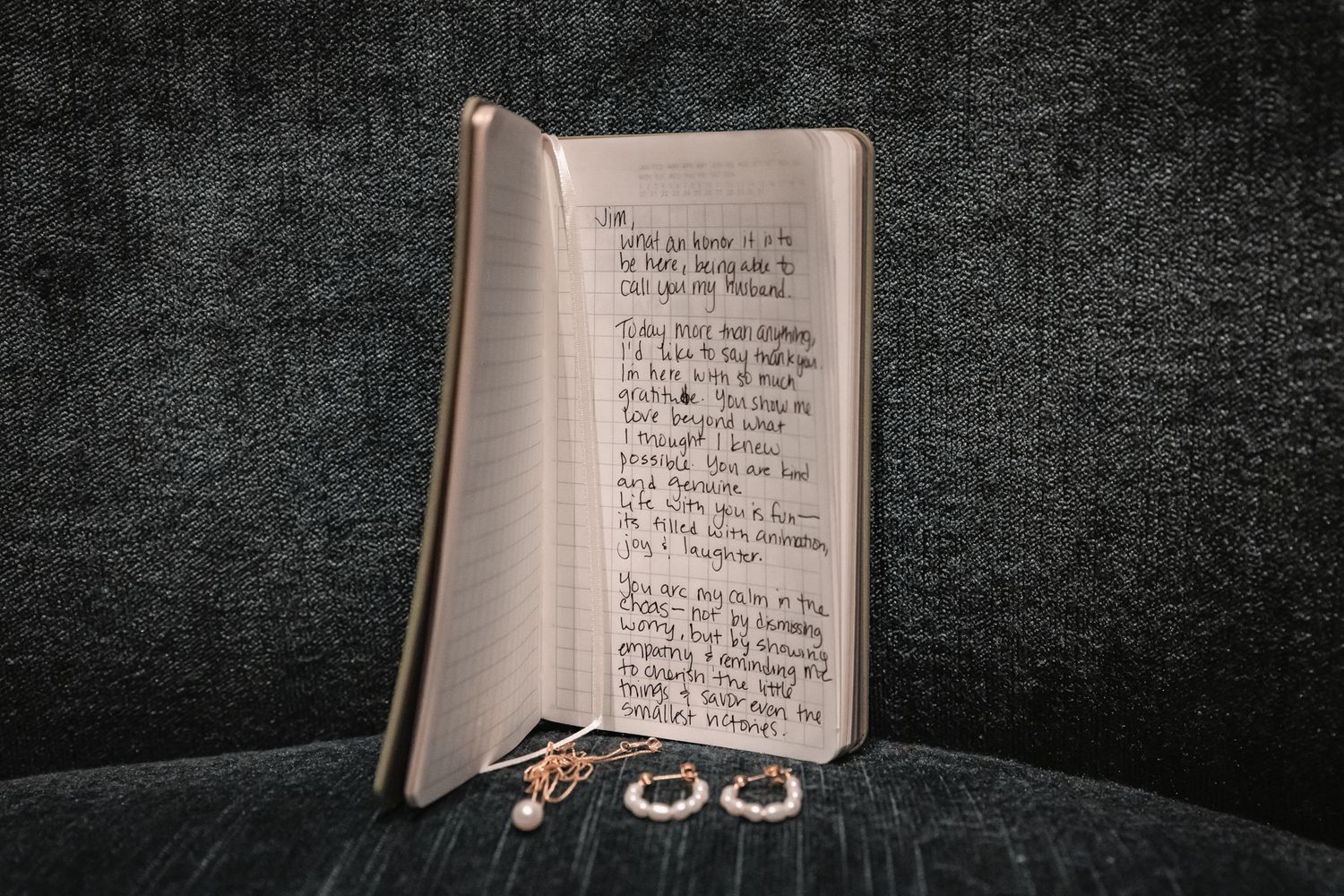 Open journal with handwritten notes beside gold jewelry on dark fabric surface.