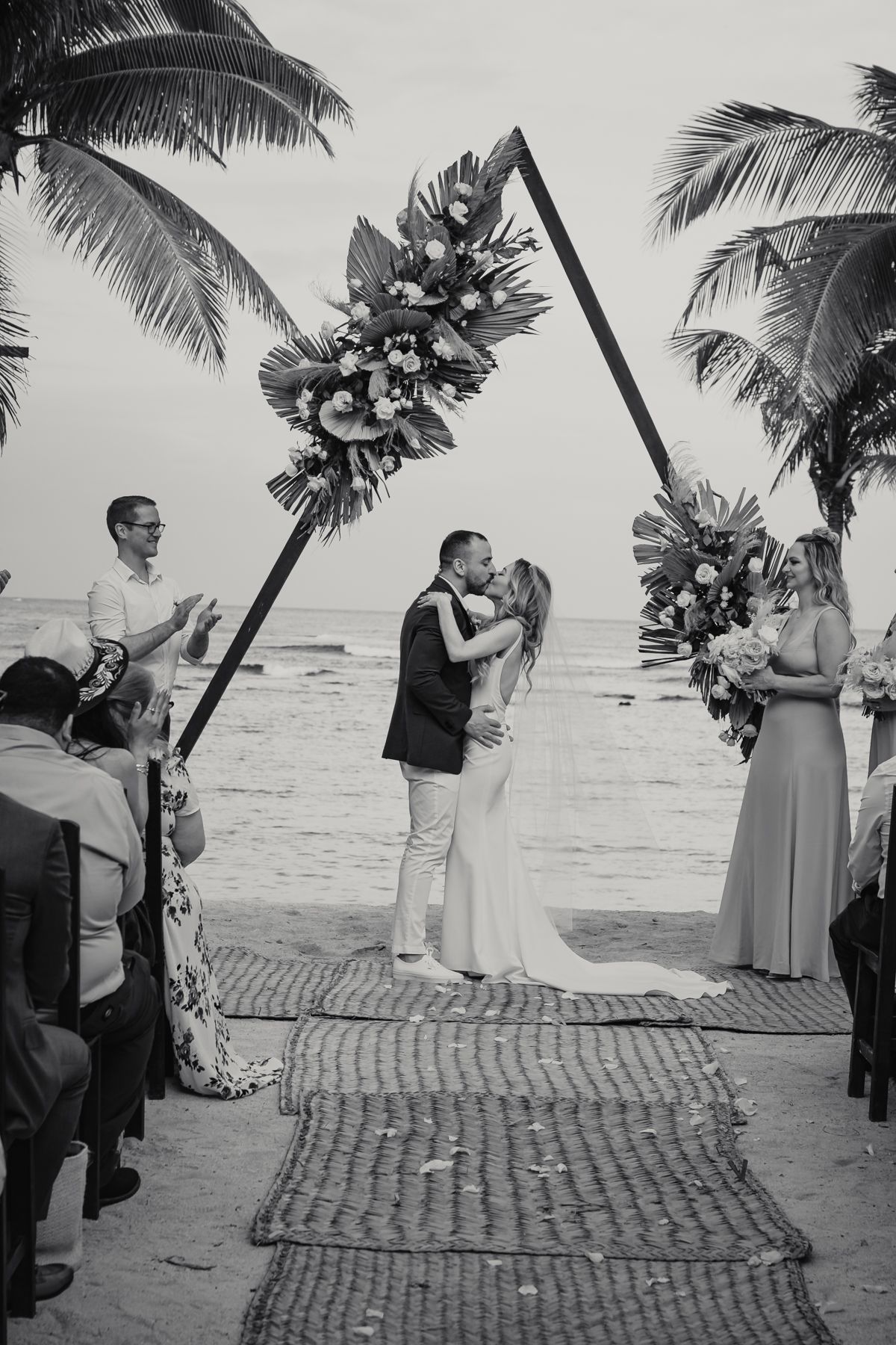 Black and white photo of bride and groom kissing under a triangular floral arch at a beach wedding.