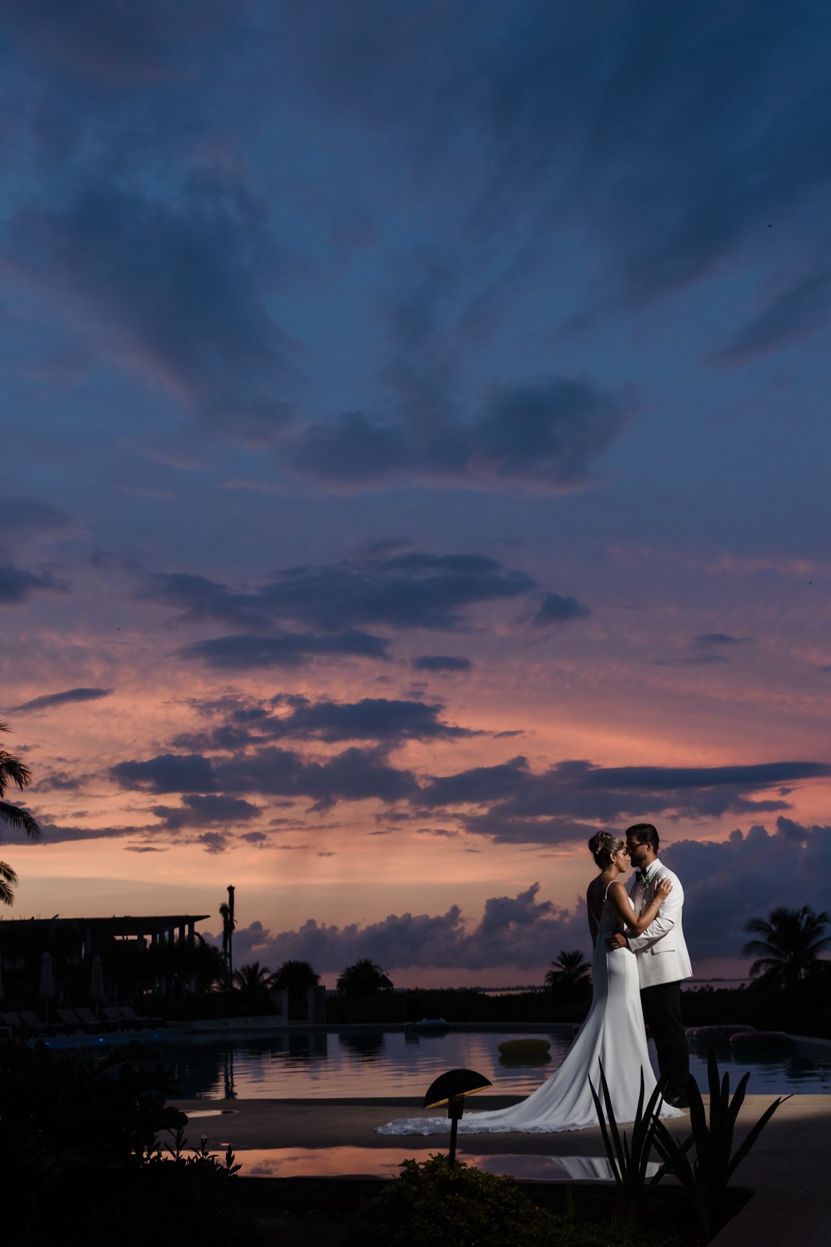 Wedding couple silhouetted against dramatic purple sunset sky beside resort infinity pool at dusk.