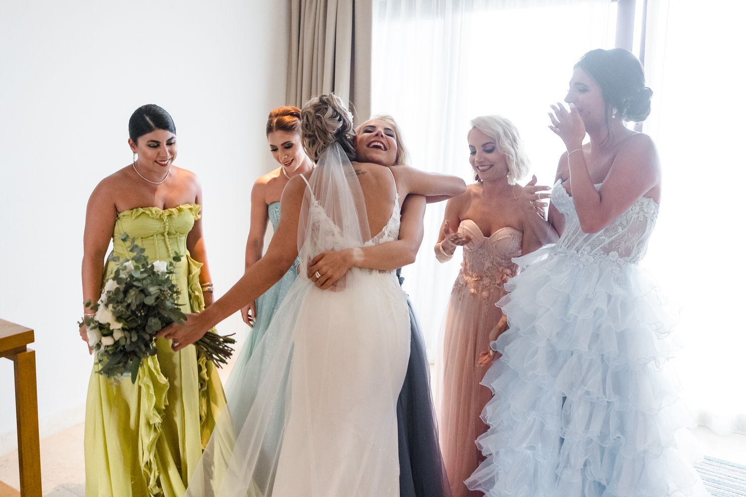 Bride hugging bridesmaid surrounded by women in colorful gowns during wedding preparations.