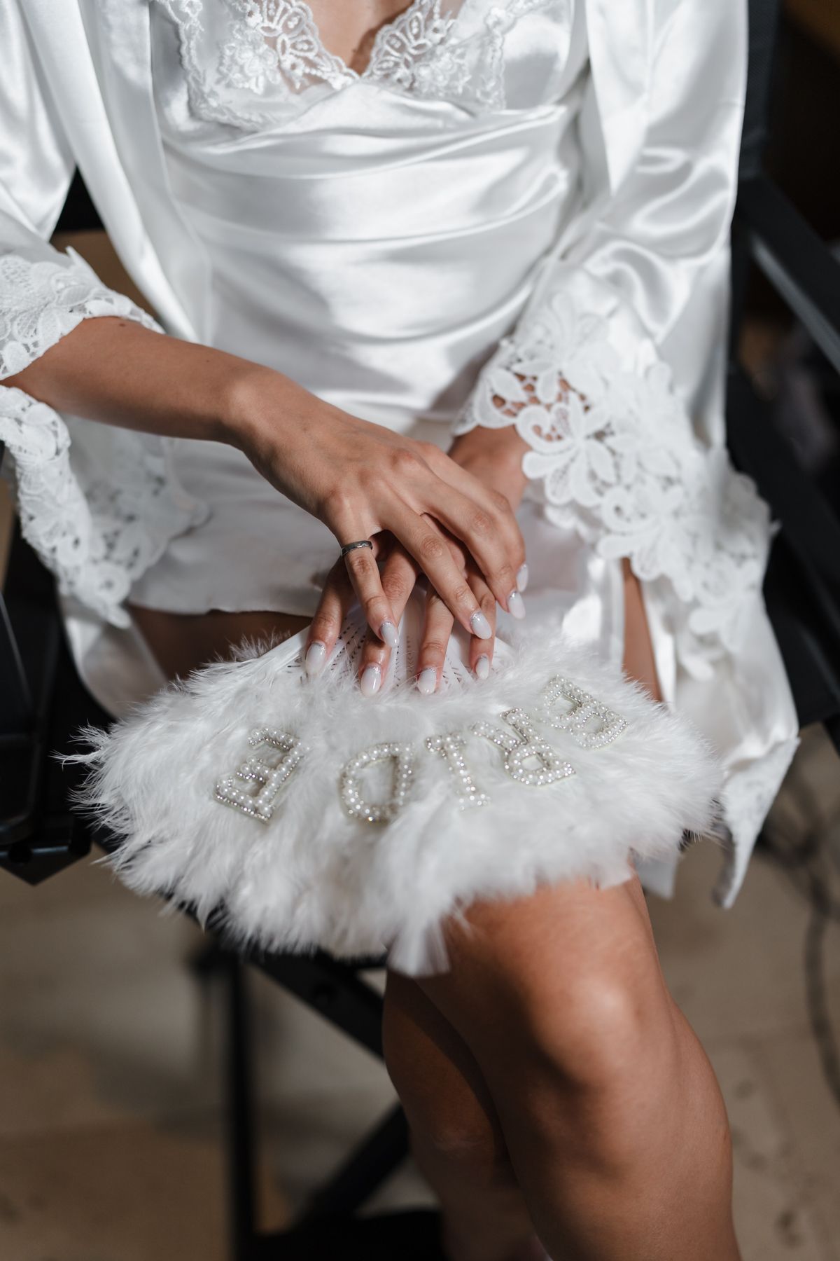 Bride in white satin lace robe holding fluffy white bridal clutch purse with pearl letters while showing engagement ring.