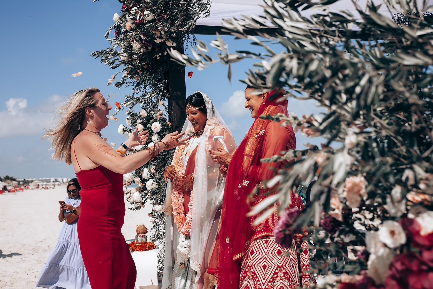 Wedding party in red dresses posing together on a sunny beach with palm trees in the background.