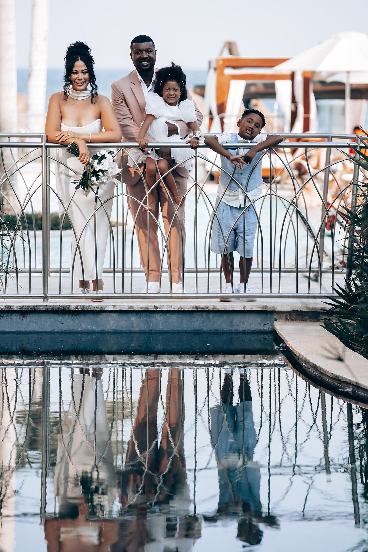 Wedding party members lean against a railing with their reflections showing in a mirrored surface below.