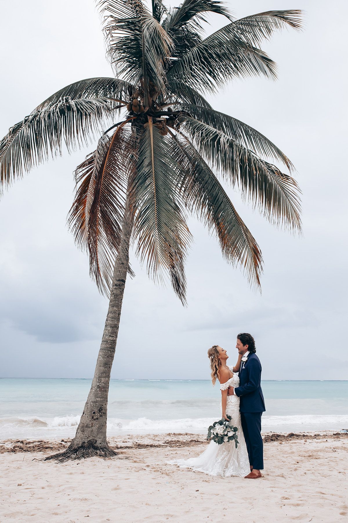 Romantic beach wedding scene with palm trees and ocean views on a cloudy day.