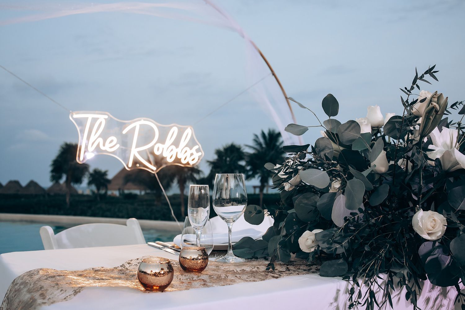 Beach wedding reception setup with string lights, floral arrangements and ocean view at sunset.