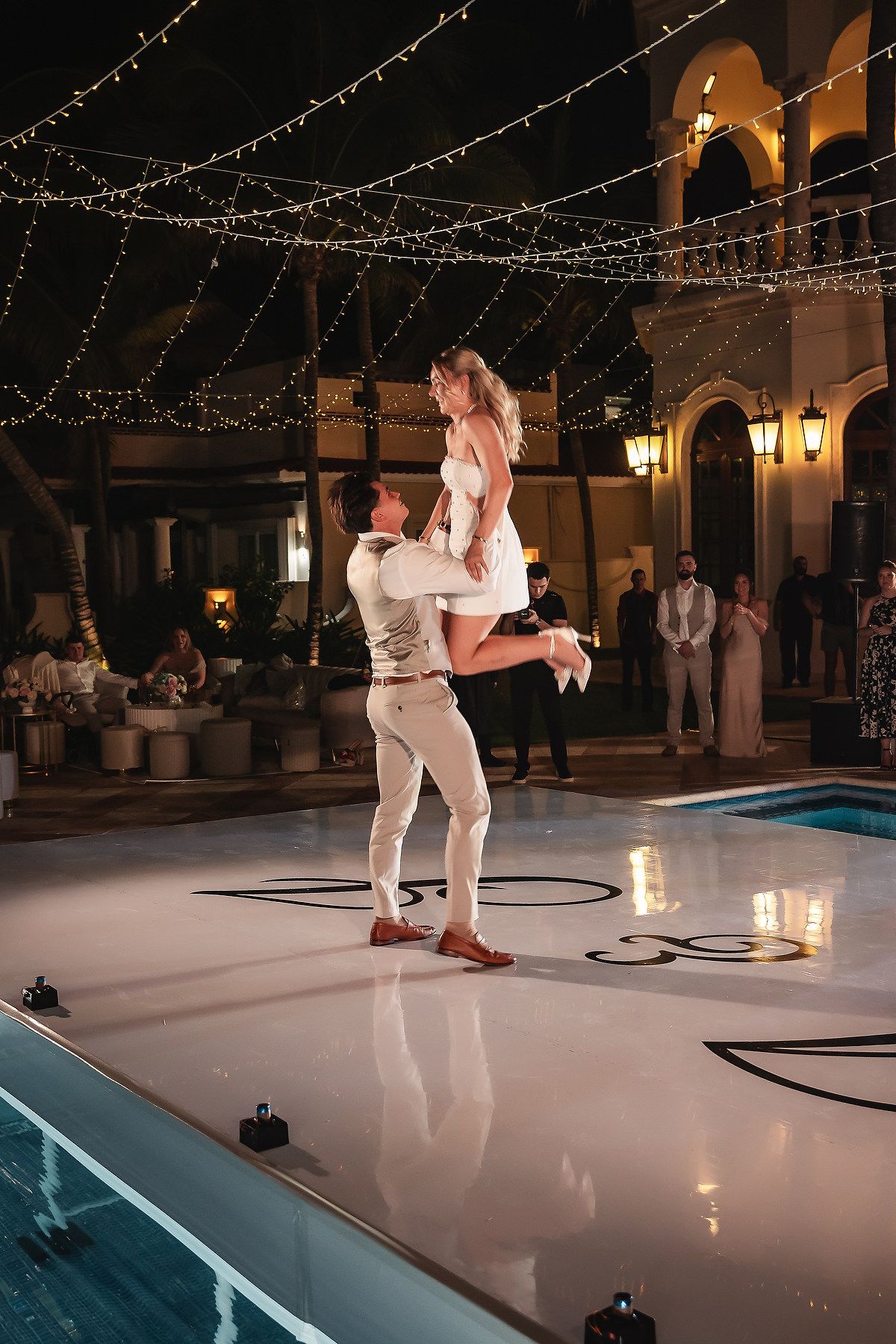 Couple dancing under string lights on a white dance floor beside a pool.