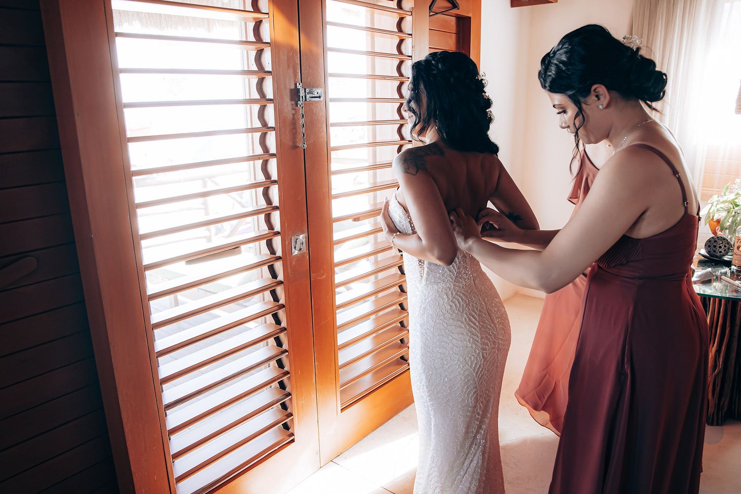Intimate wedding preparation moment captured in warm natural light through window shutters.