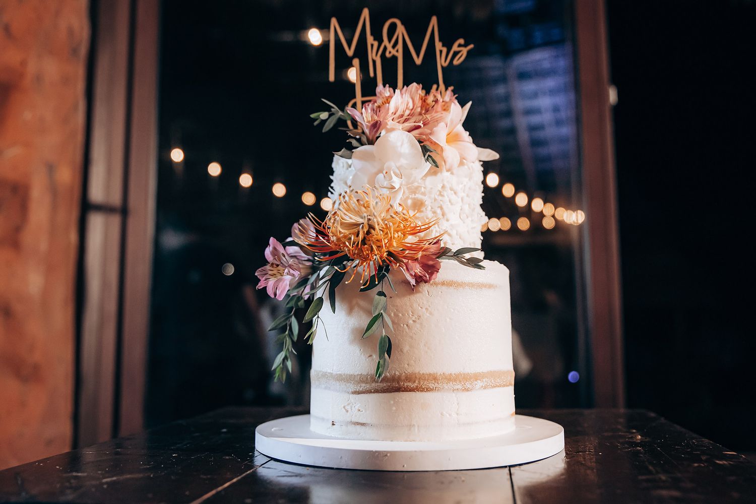 A three-tiered white wedding cake decorated with peach and coral flowers sits on a stand with string lights in background.