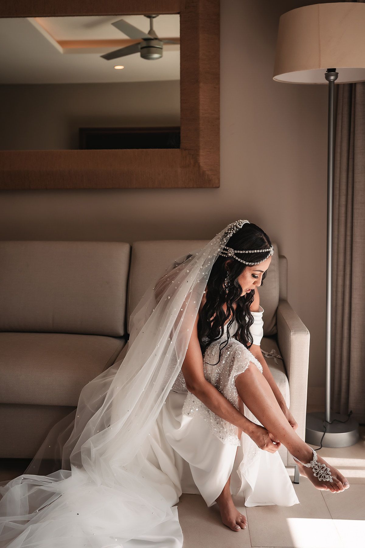 Bride in flowing white wedding dress and veil putting on shoes in an elegant indoor setting.