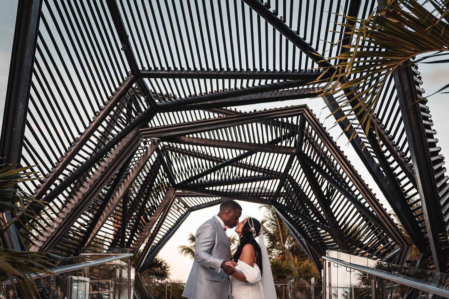 Wedding couple embracing under geometric wooden pergola structure with palm trees.