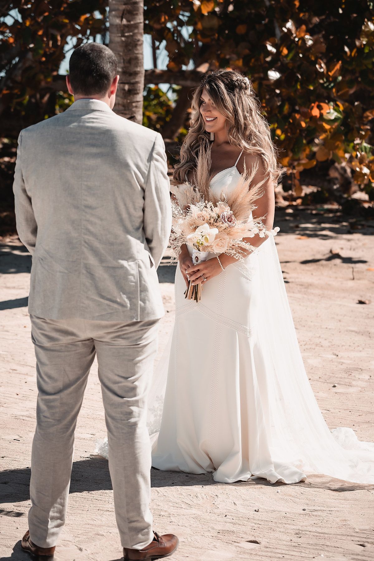 A couple in wedding attire stands together on a sunny beach path surrounded by palm trees.