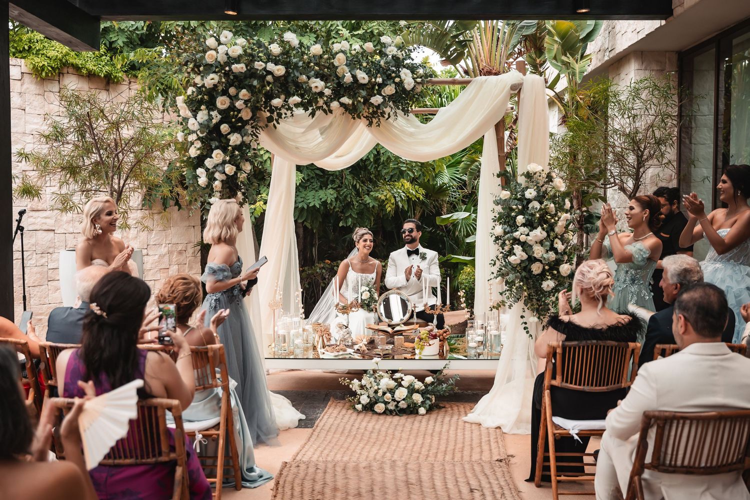 Garden wedding reception setup with wooden benches and white floral arrangements under a covered patio.