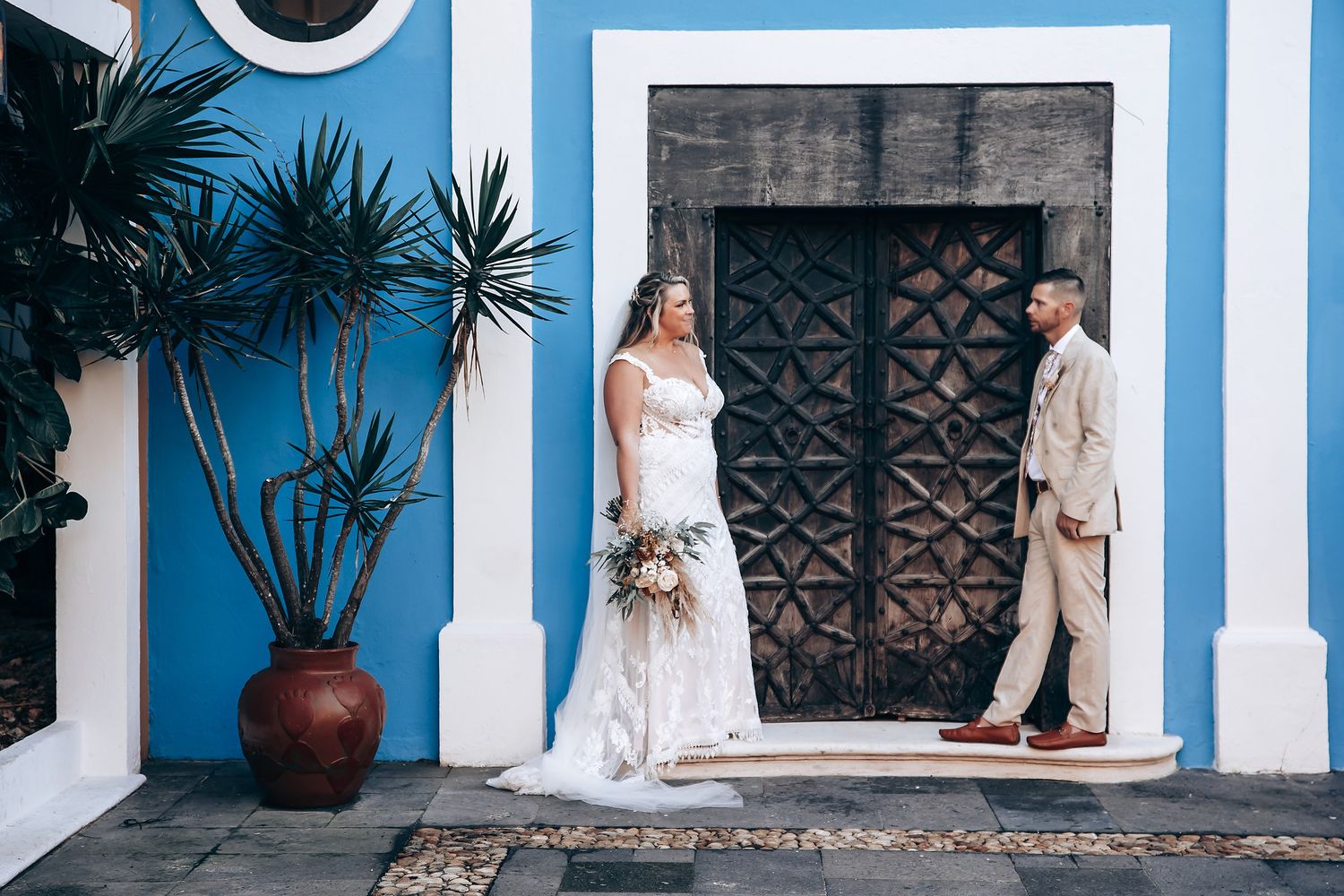 Wedding scene against blue and white colonial architecture with potted plants and ornate details.