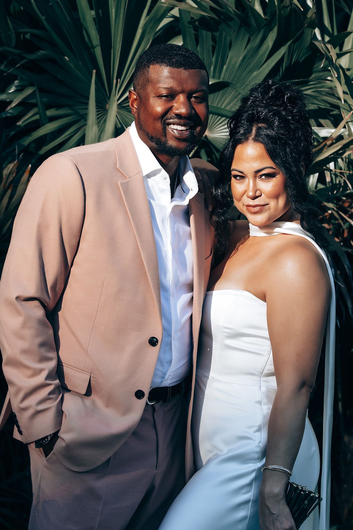 A couple poses in formal attire against tropical foliage background.