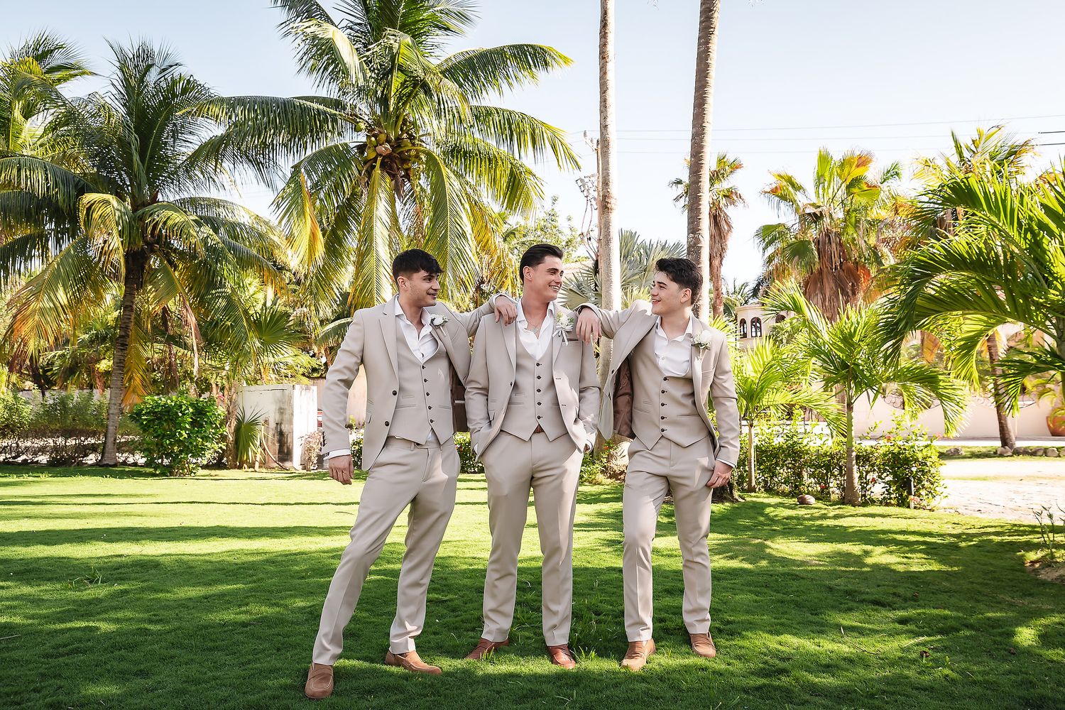 Wedding party in light gray suits poses under palm trees at a tropical beach resort.