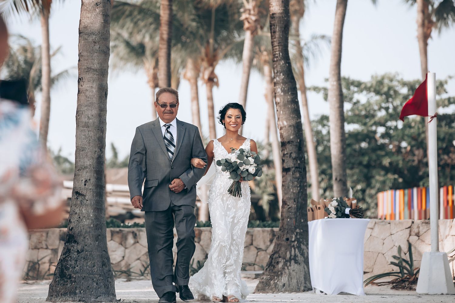 Wedding guests seated in white chairs at an outdoor tropical venue with palm trees.