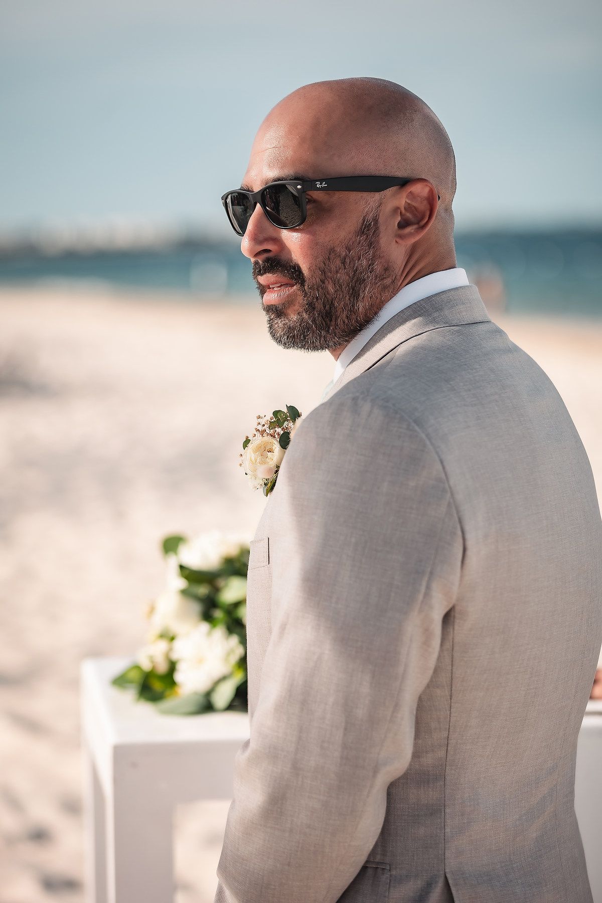 A groom in a light gray suit stands on a beach looking toward the ocean.