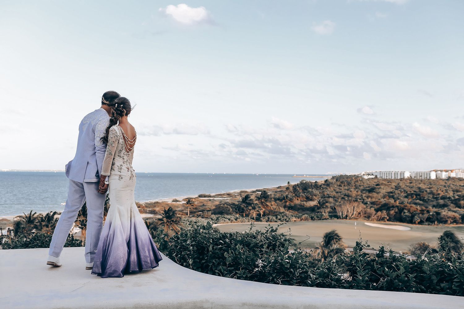 A couple in wedding attire stands together against a scenic coastal landscape during sunset.