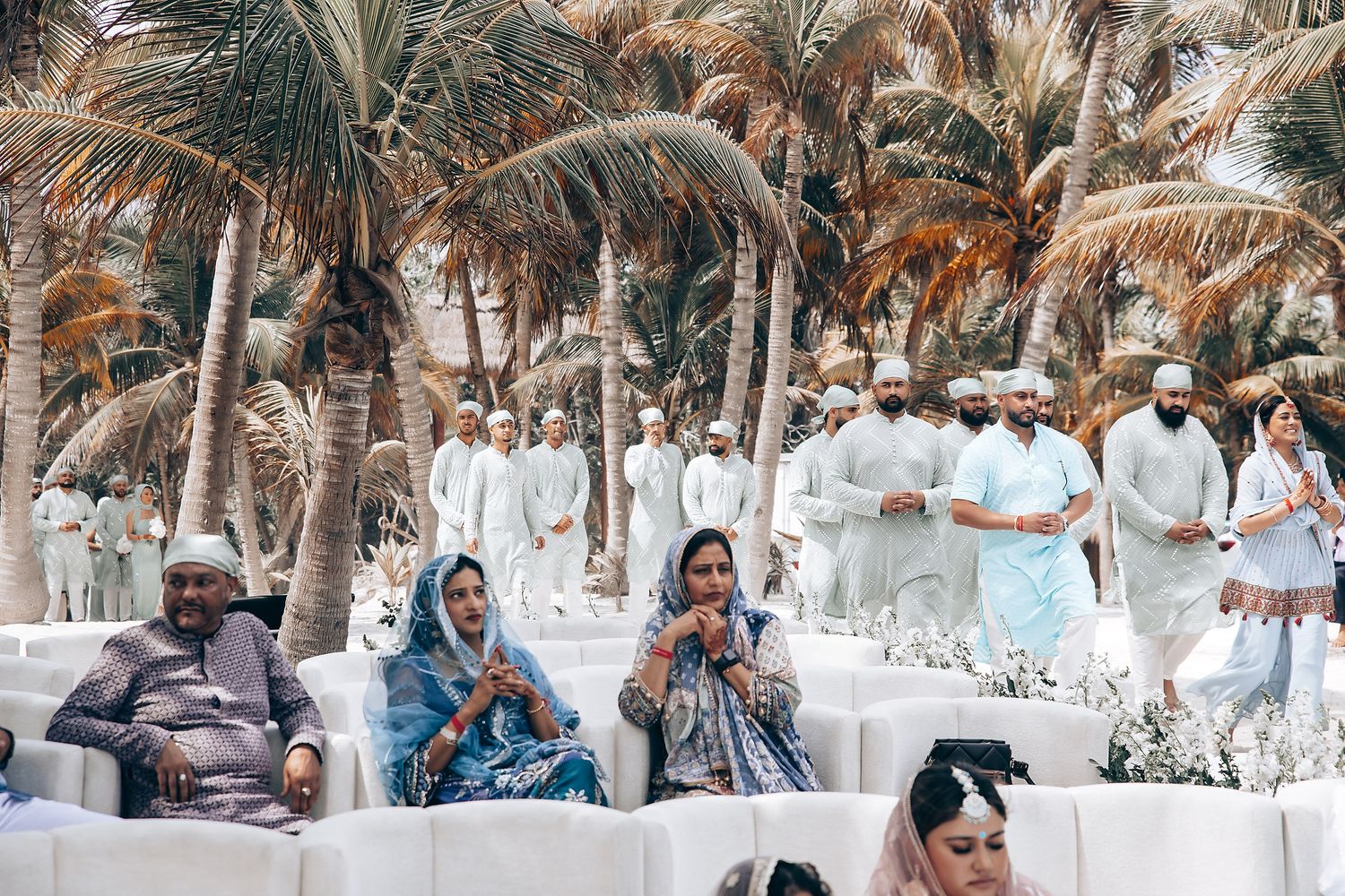A group of people in white attire gather outdoors under palm trees at a tropical beach celebration.