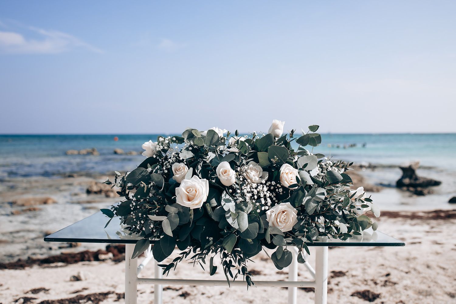 Elegant floral arrangements with white roses and greenery decorate a beachfront wedding ceremony setup.
