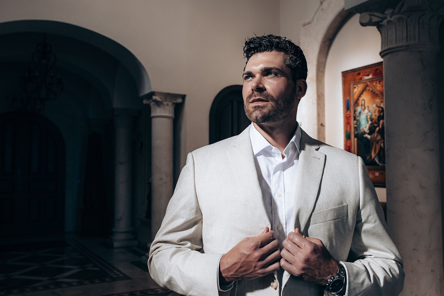 A person wearing a crisp white suit adjusts their jacket while standing in an elegant arched hallway.