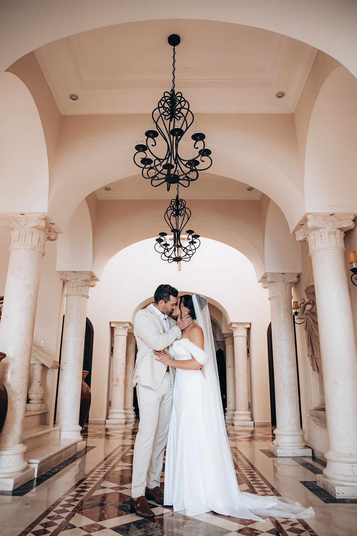 A romantic wedding couple dances beneath elegant chandeliers and white columns in a grand ballroom.