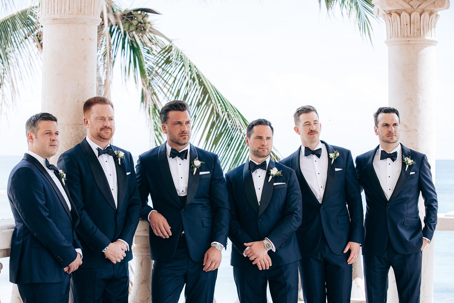Groomsmen in matching navy tuxedos and black bow ties stand together under palm trees at a tropical venue.