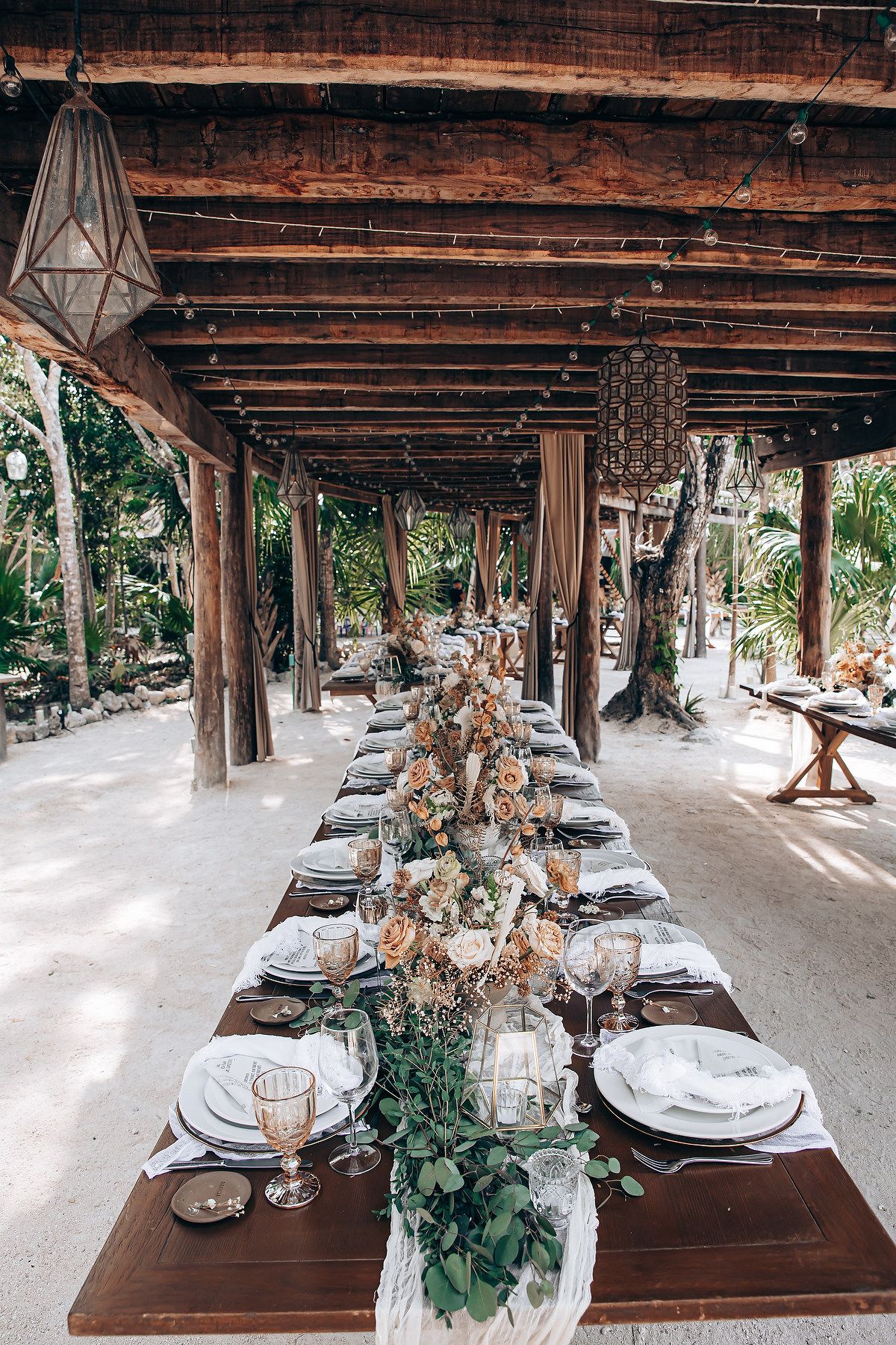 Long wooden tables set with elegant place settings under a rustic wooden pergola for an outdoor reception.