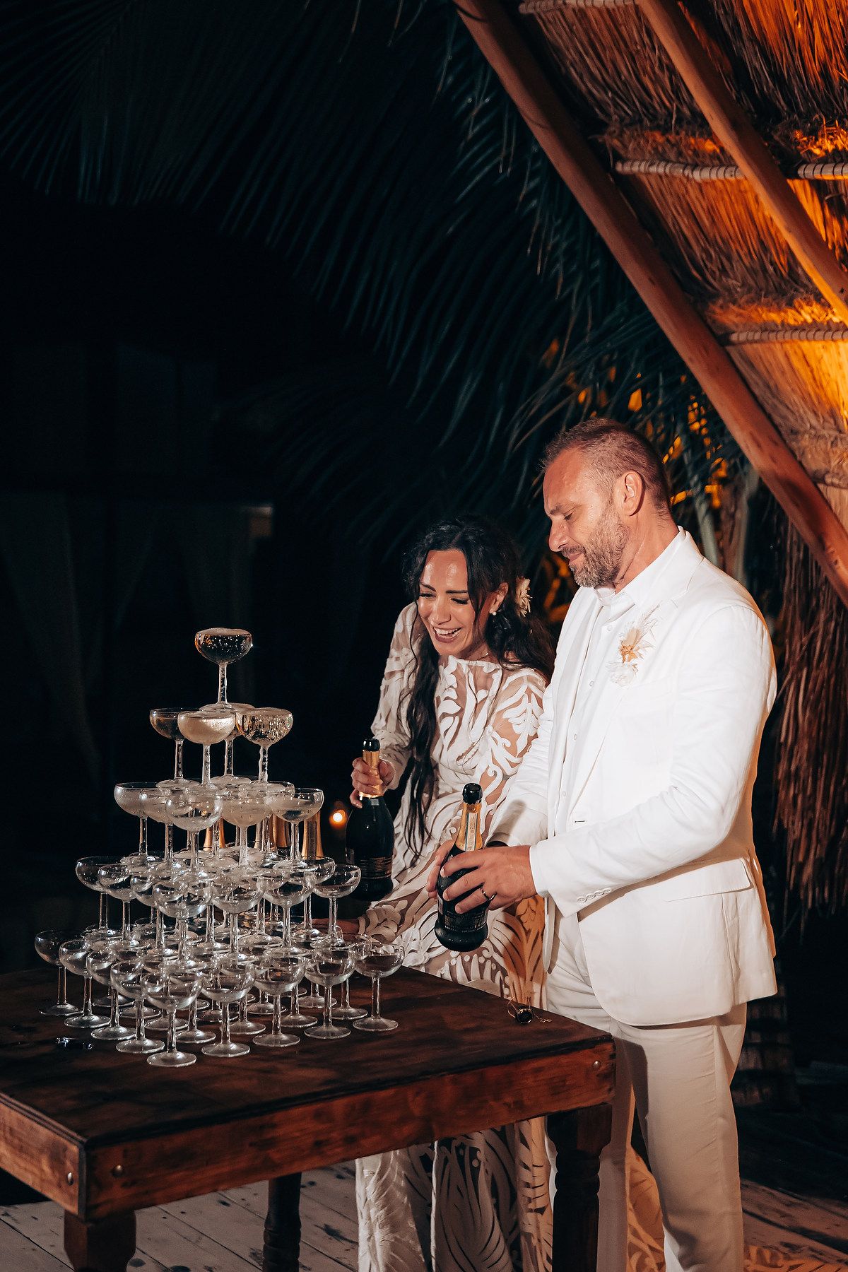 A couple pours champagne into a tower of glasses at a tropical evening celebration.