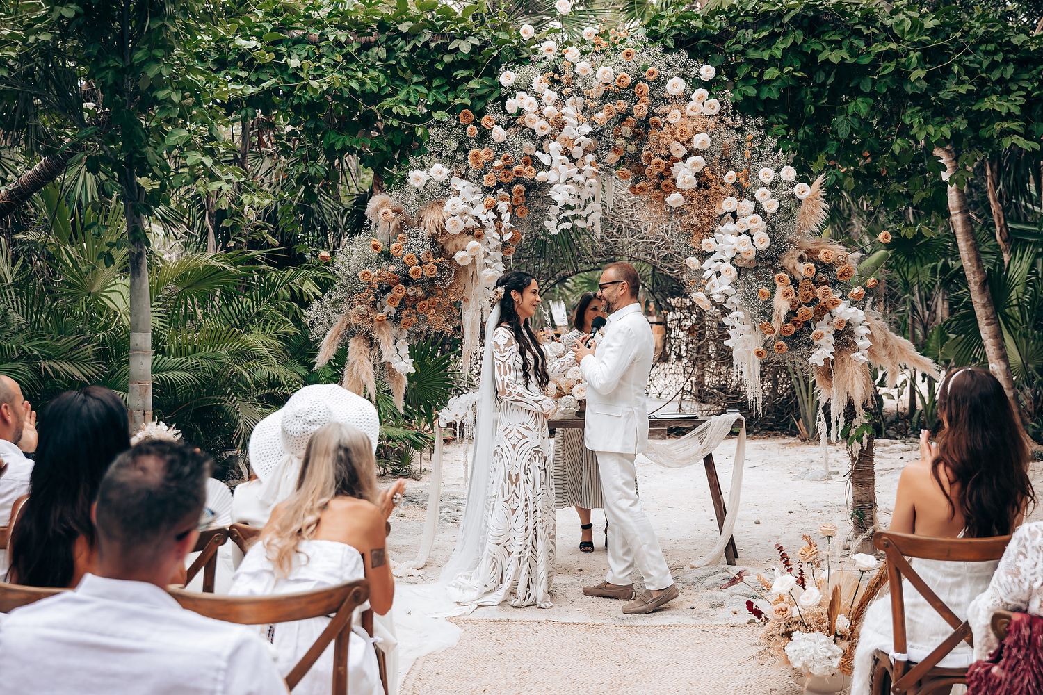 Intimate outdoor wedding ceremony with dried floral arch and wooden chairs set in a lush garden setting.