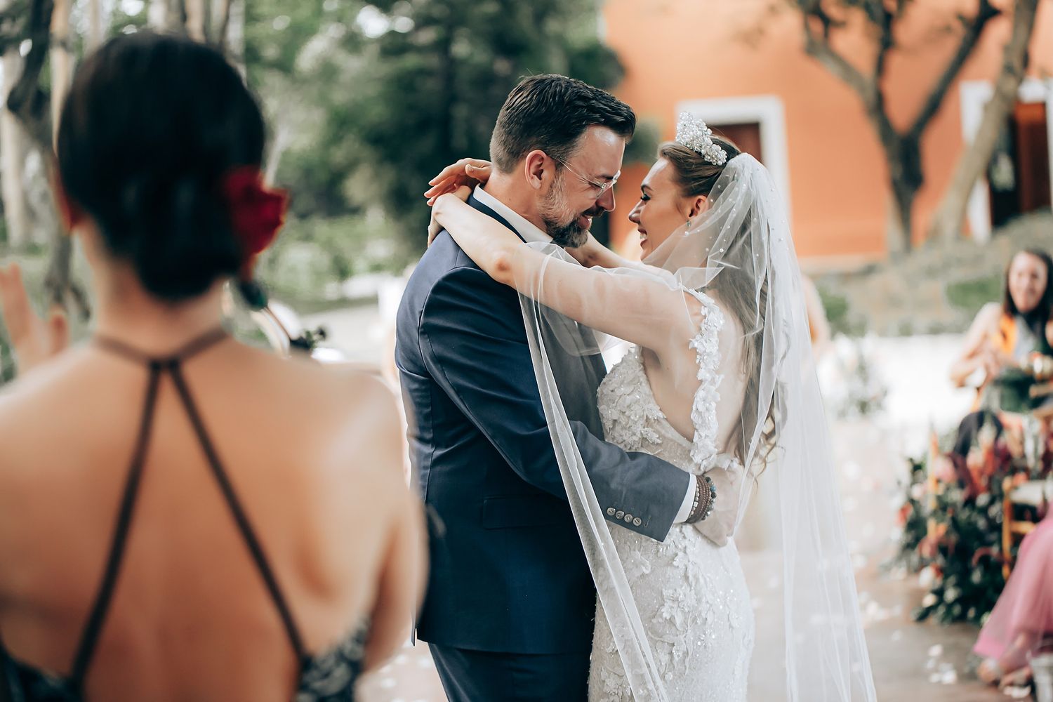 A couple shares a romantic kiss during their outdoor wedding ceremony while guests look on in the background.