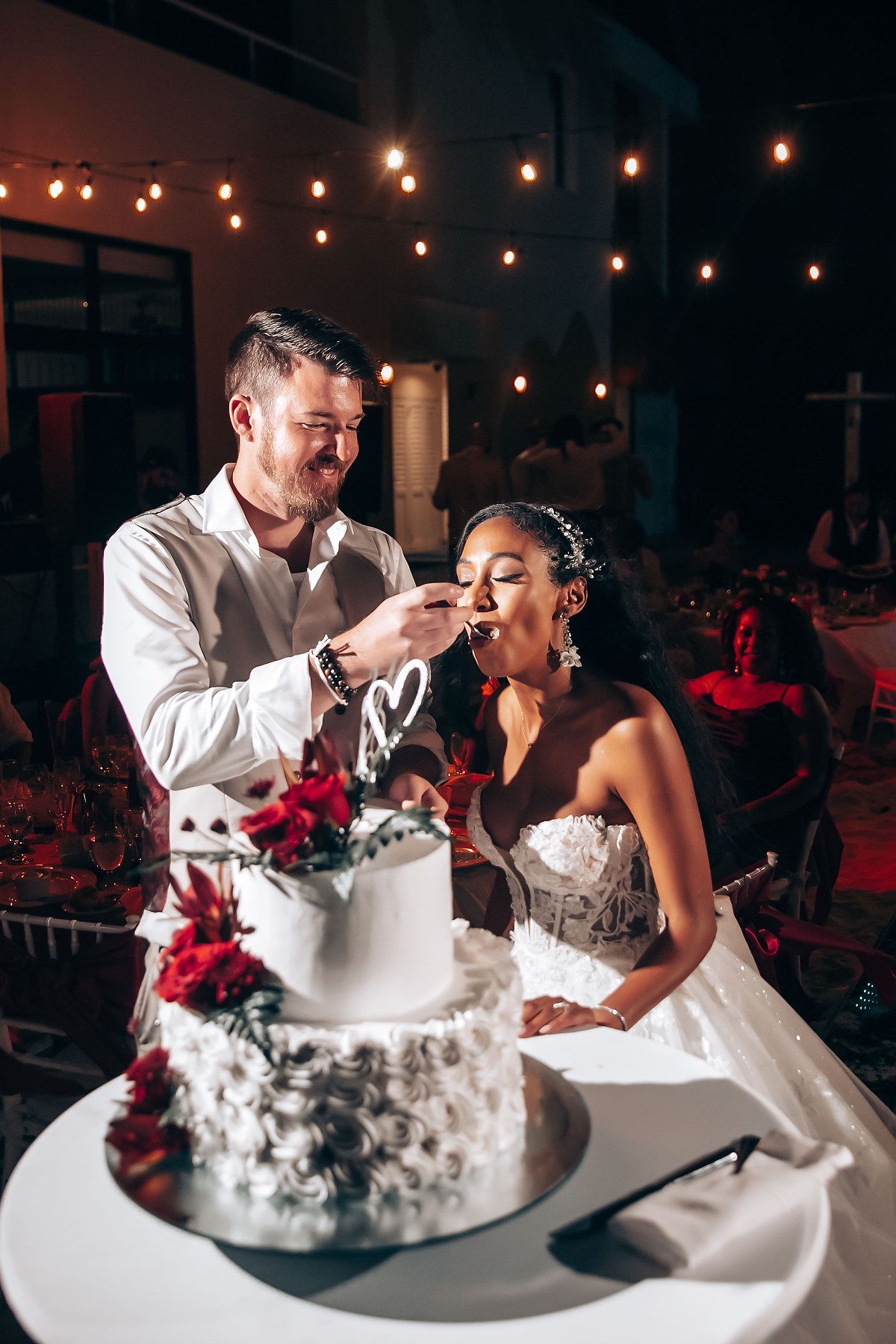 Newlyweds share cake during traditional cake feeding moment at reception under warm lighting.
