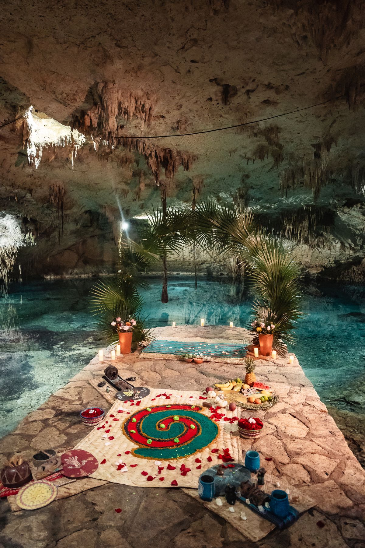 Mystical cave ceremony setup with turquoise water and red rose petals on stone platform under stalactites.